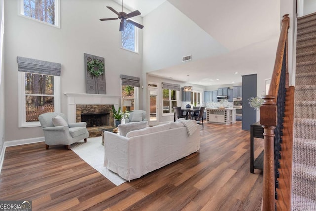 living room featuring visible vents, a ceiling fan, stairs, a stone fireplace, and dark wood-style flooring