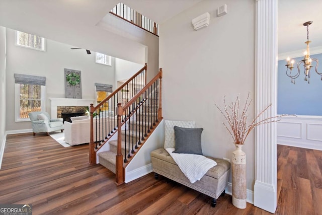 staircase with wood finished floors, baseboards, ornamental molding, a stone fireplace, and ceiling fan with notable chandelier