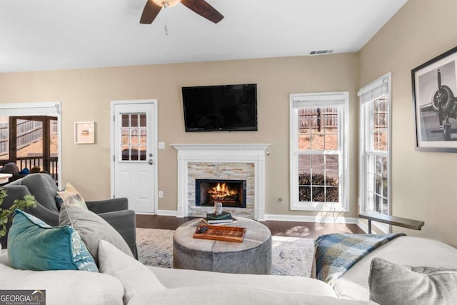 living area with visible vents, a ceiling fan, a warm lit fireplace, wood finished floors, and baseboards