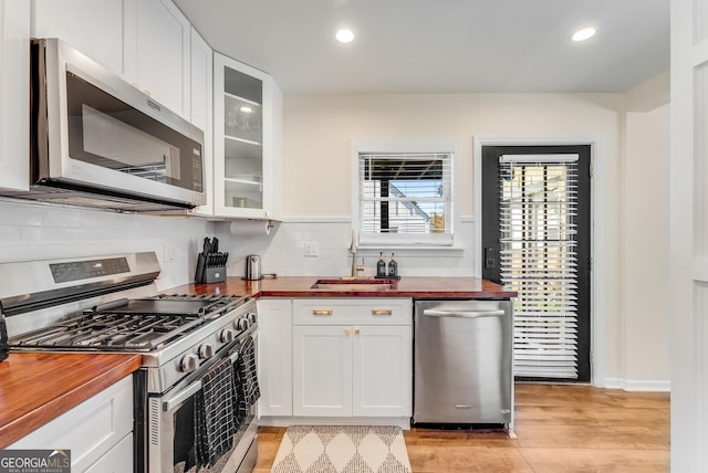 kitchen featuring glass insert cabinets, butcher block counters, stainless steel appliances, white cabinetry, and a sink