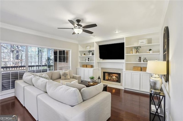 living room featuring built in shelves, crown molding, a fireplace, a ceiling fan, and dark wood-style flooring