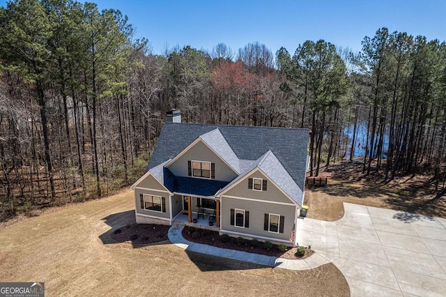 view of front of property featuring a wooded view, a porch, a shingled roof, a chimney, and concrete driveway