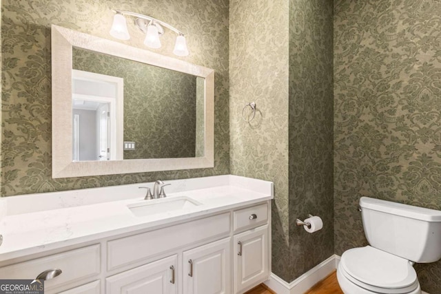 half bathroom featuring toilet, wood finished floors, wallpapered walls, baseboards, and vanity