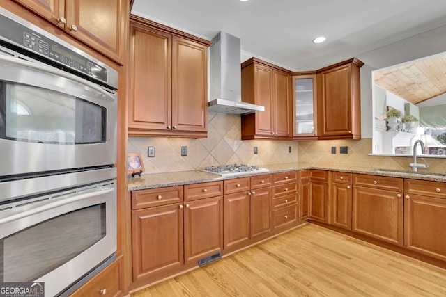 kitchen with light wood-type flooring, a sink, stainless steel double oven, wall chimney exhaust hood, and brown cabinetry