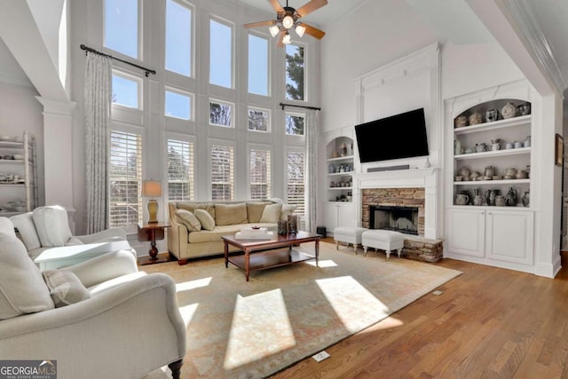 living room featuring built in features, light wood-type flooring, a stone fireplace, a ceiling fan, and ornate columns