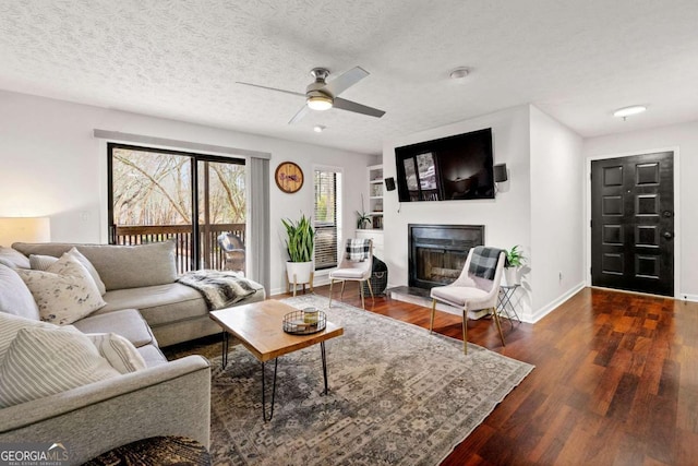 living room with a ceiling fan, a textured ceiling, a glass covered fireplace, wood finished floors, and baseboards