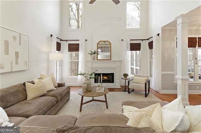 living room featuring a ceiling fan, wood finished floors, ornate columns, a fireplace with flush hearth, and a towering ceiling