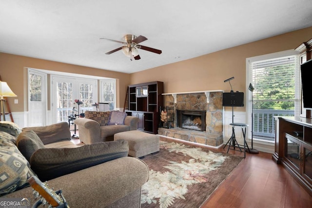 living area with a stone fireplace, wood finished floors, a wainscoted wall, and ceiling fan