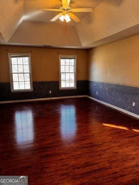 empty room with dark wood-style floors, a wainscoted wall, lofted ceiling, ceiling fan, and a raised ceiling