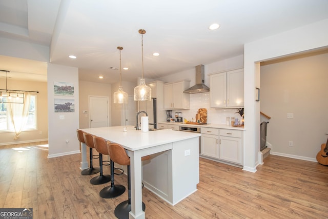 kitchen featuring a kitchen bar, a center island with sink, light wood-style flooring, wall chimney exhaust hood, and light countertops