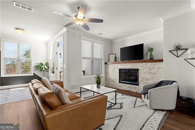 living room featuring visible vents, ceiling fan, ornamental molding, a stone fireplace, and wood finished floors