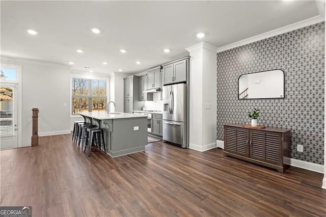 kitchen featuring wallpapered walls, an island with sink, gray cabinetry, stainless steel appliances, and crown molding