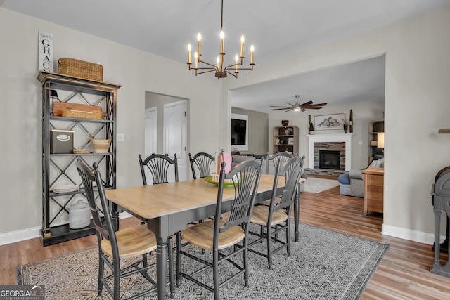 dining room with baseboards, light wood-style floors, a stone fireplace, and ceiling fan with notable chandelier