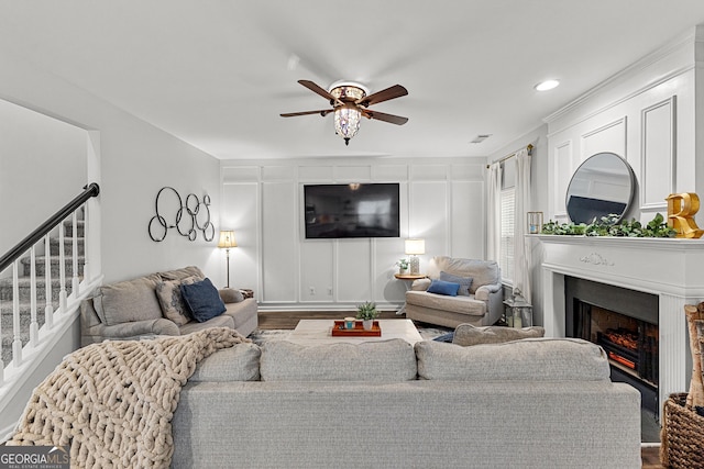 living area featuring visible vents, a warm lit fireplace, ceiling fan, stairs, and a decorative wall