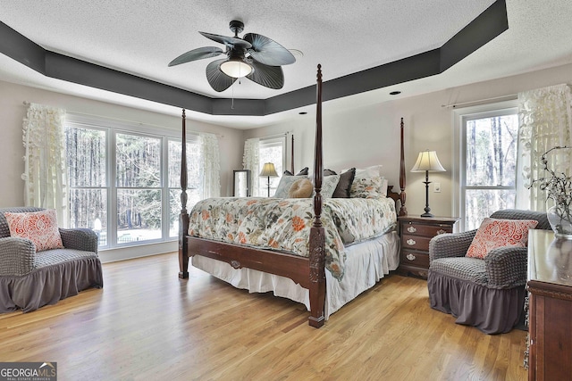 bedroom with a tray ceiling, multiple windows, a textured ceiling, and light wood-style flooring