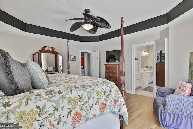 bedroom with light wood finished floors, connected bathroom, ceiling fan, a textured ceiling, and a raised ceiling