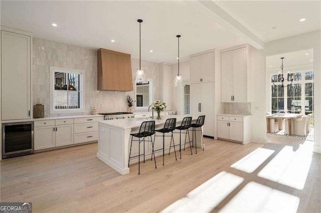 kitchen with beverage cooler, custom exhaust hood, light wood-style flooring, a kitchen bar, and backsplash