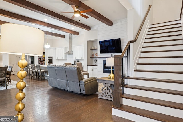 living room featuring ceiling fan, stairway, beamed ceiling, a fireplace, and dark wood-style floors