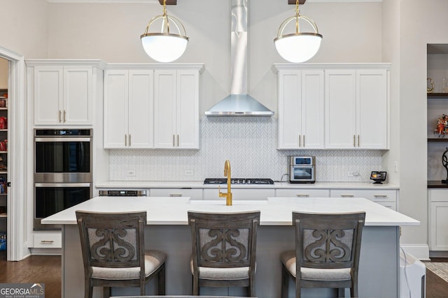 kitchen with tasteful backsplash, wall chimney exhaust hood, stainless steel double oven, gas cooktop, and a kitchen island with sink