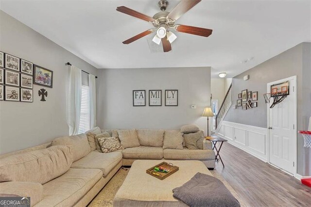 living room featuring ceiling fan, stairway, wainscoting, wood finished floors, and a decorative wall