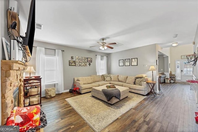 living area with visible vents, a stone fireplace, dark wood-type flooring, and a ceiling fan