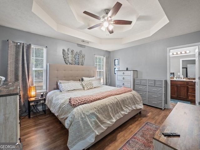 bedroom with dark wood-style floors, ensuite bath, a tray ceiling, ceiling fan, and a textured ceiling