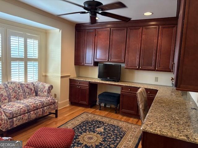 kitchen with light stone counters, light wood finished floors, ornamental molding, ceiling fan, and built in desk