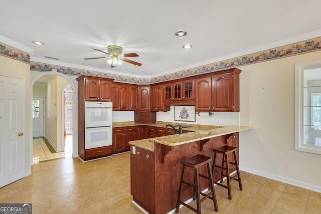 kitchen featuring a kitchen bar, arched walkways, double oven, a peninsula, and light stone countertops