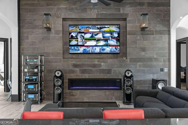 living area with arched walkways, a glass covered fireplace, a ceiling fan, and wood finished floors