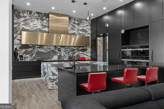 kitchen featuring light wood finished floors, a breakfast bar area, dark stone countertops, range hood, and dark cabinetry