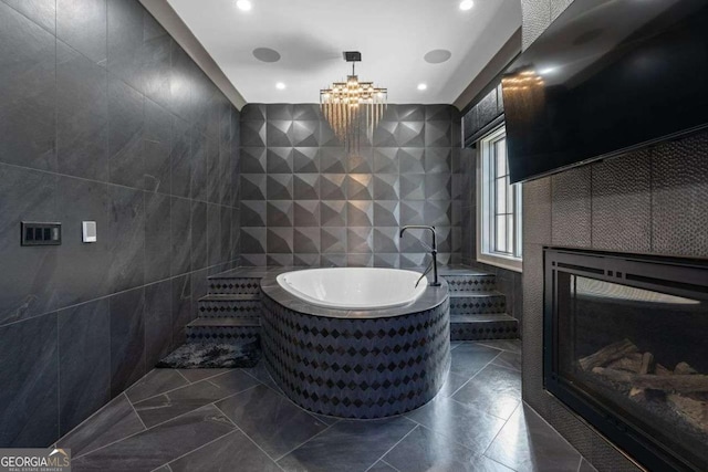 bathroom featuring a garden tub, a notable chandelier, tile walls, and a glass covered fireplace