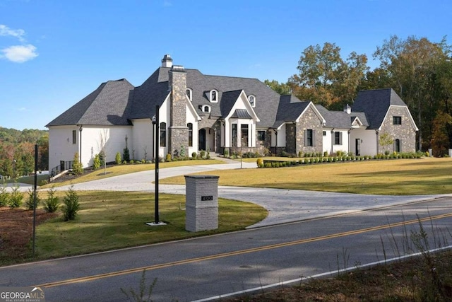 french country home featuring stone siding, curved driveway, a chimney, and a front lawn