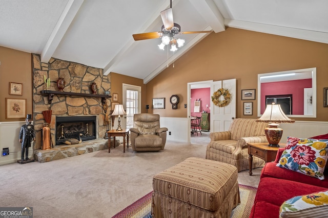 living area featuring carpet, lofted ceiling with beams, ceiling fan, a stone fireplace, and wainscoting