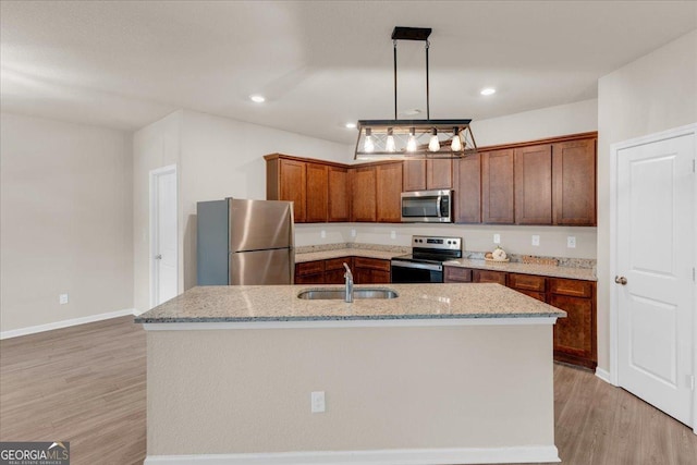 kitchen with a kitchen island with sink, a sink, light wood-style floors, appliances with stainless steel finishes, and light stone countertops