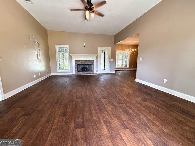 unfurnished living room with a wealth of natural light, a stone fireplace, dark wood-style floors, and ceiling fan with notable chandelier