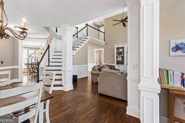 living room featuring a wainscoted wall, dark wood-type flooring, ceiling fan with notable chandelier, stairway, and ornate columns