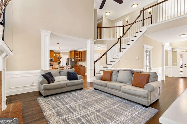 living area with stairs, decorative columns, ceiling fan with notable chandelier, a high ceiling, and dark wood-style floors