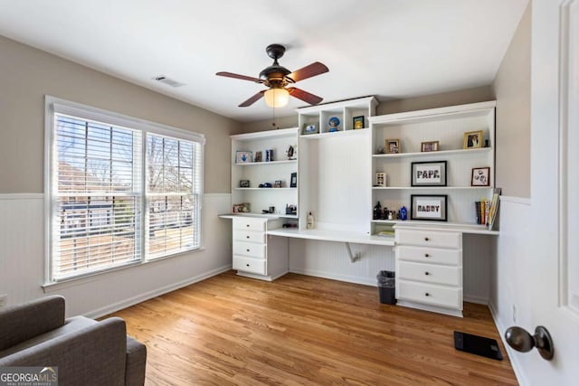 home office with light wood finished floors, visible vents, wainscoting, built in study area, and a ceiling fan