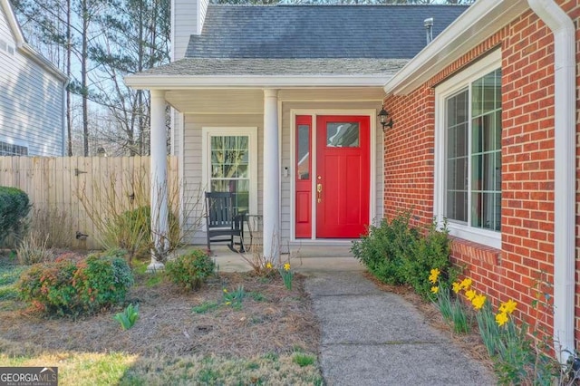 view of exterior entry with brick siding, a shingled roof, a chimney, and fence