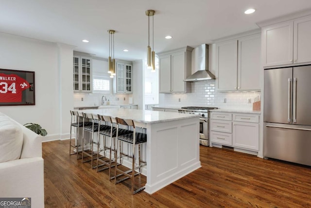 kitchen featuring high quality appliances, a breakfast bar area, wall chimney range hood, glass insert cabinets, and dark wood-style flooring