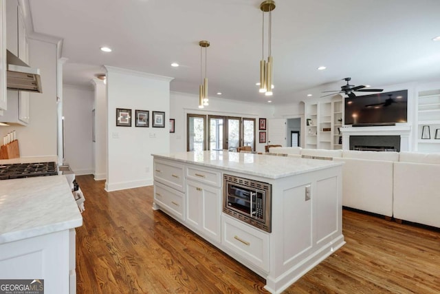 kitchen featuring wood finished floors, a fireplace, white cabinets, under cabinet range hood, and stainless steel microwave