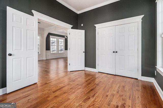 unfurnished bedroom with baseboards, light wood finished floors, arched walkways, a closet, and crown molding