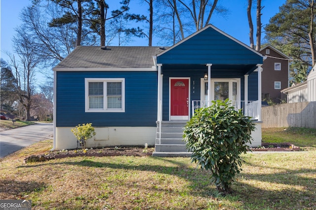 bungalow-style home with covered porch, a shingled roof, a front lawn, and fence