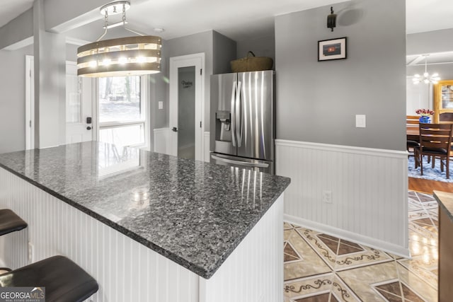 kitchen with light tile patterned floors, dark stone counters, wainscoting, a notable chandelier, and stainless steel fridge