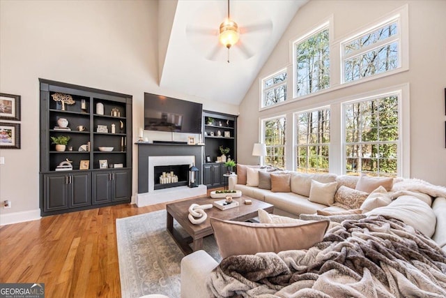 living area with a ceiling fan, baseboards, light wood finished floors, high vaulted ceiling, and a fireplace with raised hearth