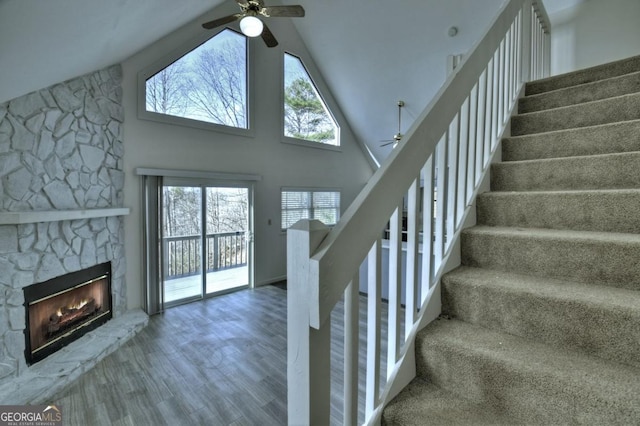 staircase featuring a fireplace, high vaulted ceiling, a ceiling fan, and wood finished floors