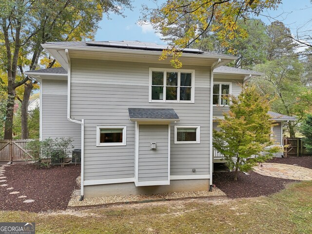 rear view of property featuring cooling unit, roof mounted solar panels, roof with shingles, and fence