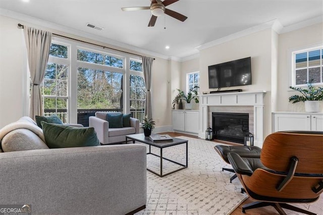 living room featuring visible vents, ornamental molding, a glass covered fireplace, baseboards, and ceiling fan