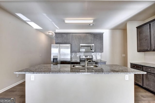 kitchen featuring a kitchen island with sink, a sink, light stone counters, stainless steel appliances, and dark brown cabinetry