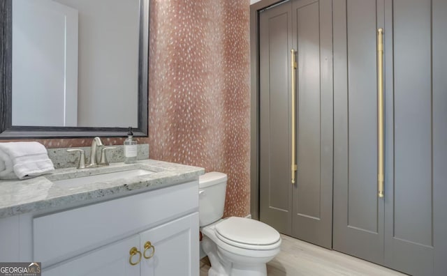 half bathroom with toilet, wood finished floors, vanity, and wallpapered walls